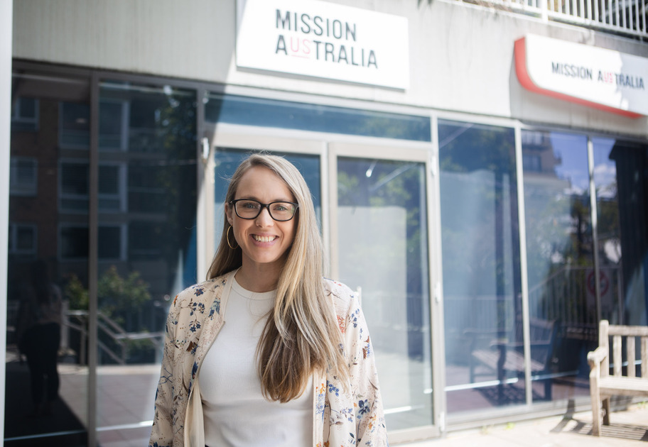 A woman with long blonde hair and glasses, wearing a floral blazer and white top, stands smiling in front of a Mission Australia office building.
