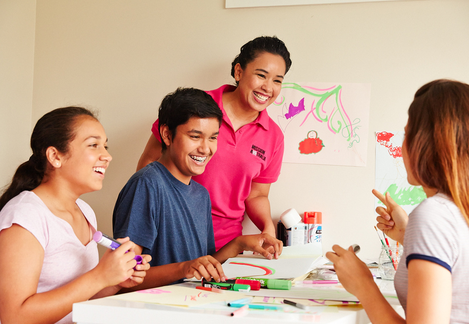 Three teens sit around a table, smiling and laughing while doing arts and crafts a Mission Australia case worker stands next to them. Drawings and art supplies are scattered on the table and colorful artwork is displayed on the wall behind them.