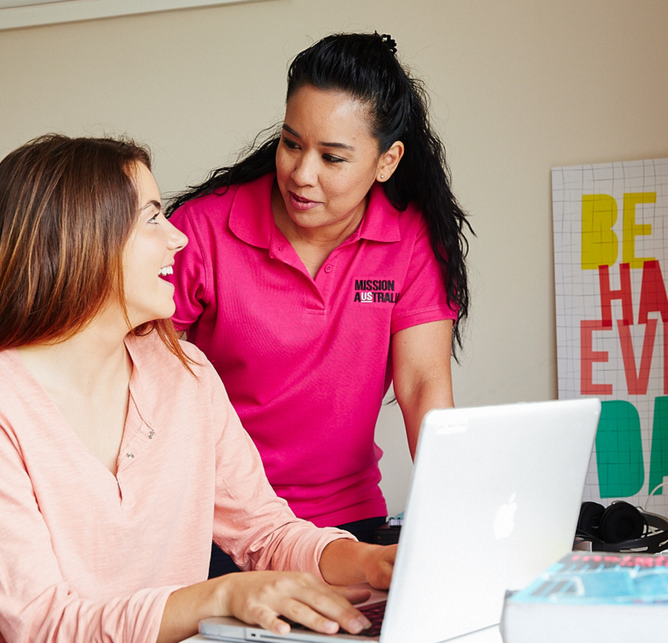 Two women are smiling and talking at a desk with a laptop. One is seated and typing, while the other, a Mission Australia case worker, stands beside her.  