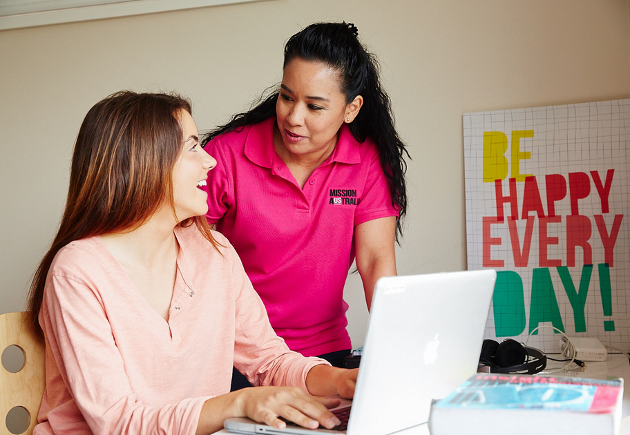 Two women are smiling and talking at a desk with a laptop. One is seated and typing, while the other, a Mission Australia case worker, stands beside her.  