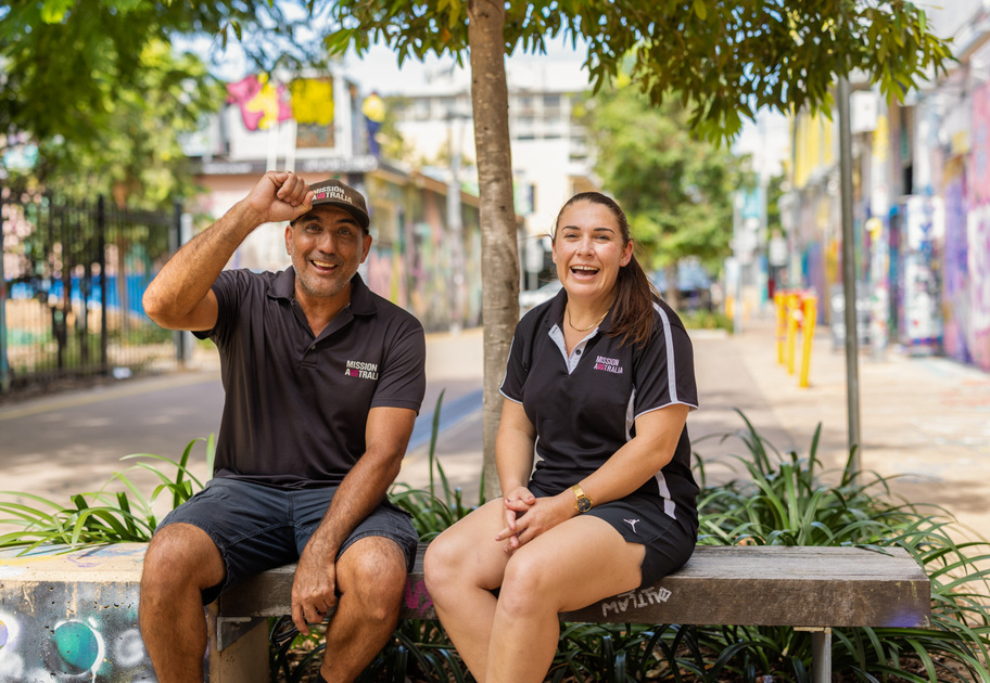 Billy Kruse and Megan Dennis from Mission Australia wear matching Mission Australia black shirts. They sit on a bench outdoors, smiling and laughing. The man raises his cap in greeting. Colorful graffiti walls and greenery are visible in the background.
