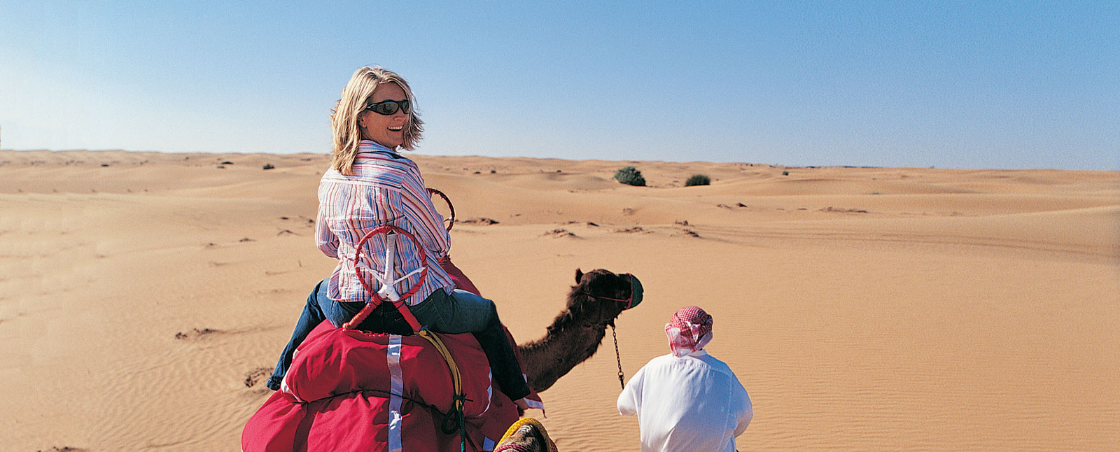 A female solo traveler smiles back at the camera while riding a camel through a vast desert with a guide leading the way.