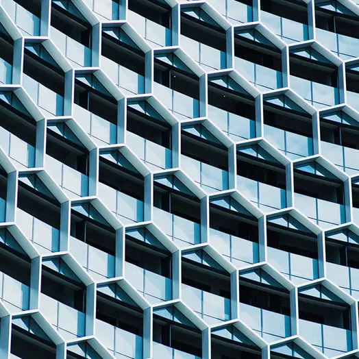 
A close-up architectural shot of a modern building's facade, featuring a repeating pattern of light-colored, projecting hexagonal window frames filled with reflective blue-tinted glass. The design creates a deep, three-dimensional geometric grid.
