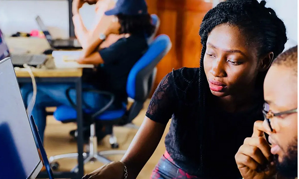 A woman in a black top points to a monitor while collaborating closely with a man wearing glasses in an office setting. Another person is blurred in the background.