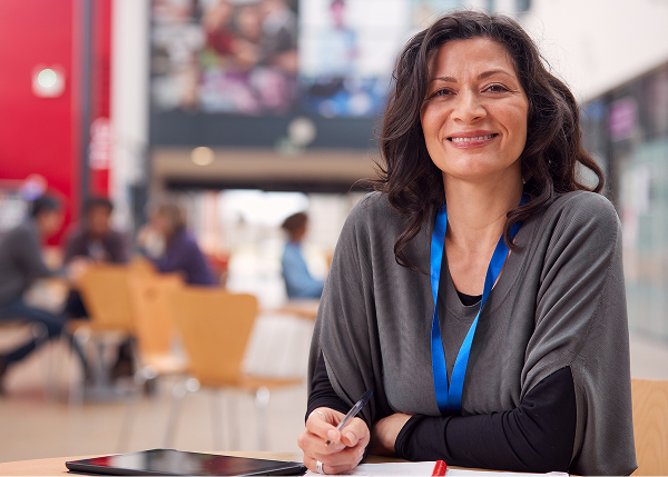 A smiling woman with dark, wavy hair and a gray top sits at a desk, pen in hand, with a tablet in front of her. She is wearing a blue lanyard, and a busy, blurred communal area is behind her.