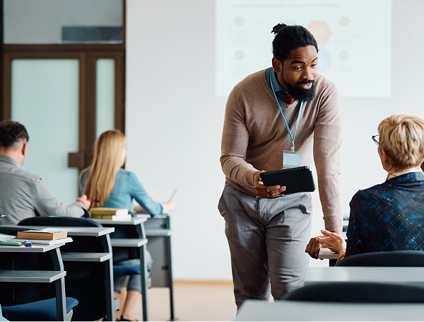 A Black male instructor with a beard and hair pulled back, wearing a light brown sweater and a lanyard, stands and leans toward a student, talking to them in a classroom. Other students are seated at desks in the background.