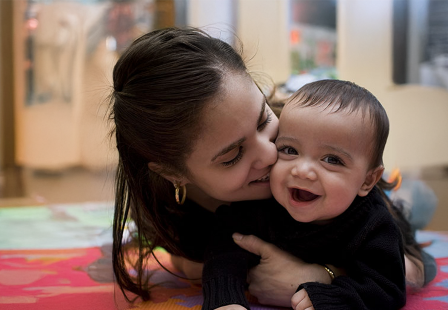 A smiling mother lying on a colorful play mat with her happy baby, engaging in a playful and affectionate moment.