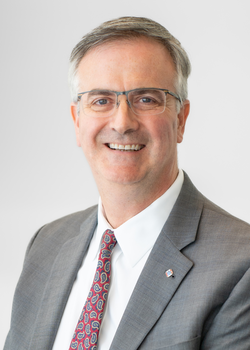 A corporate headshot style portrait of Thomas Burnford, D.Min., smiling wearing a light grey suit, white shirt, and red patterned tie on a light grey background.