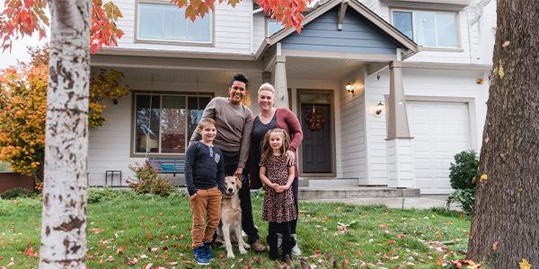 An interracial lesbian couple stands smiling with their two kids in front of their new home