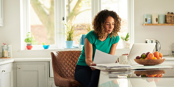 A woman with brown skin and curly hair stands at a kitchen island going over paperwork in front of her laptop