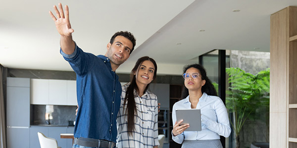 A man and woman stand smiling in a home while they talk to a woman holding a tablet