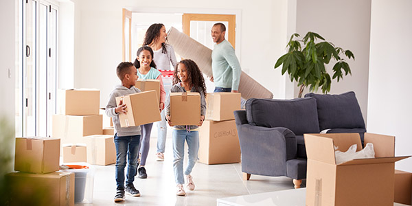 A Black man and Black woman smile at each other as they carry items into a home with their three laughing children