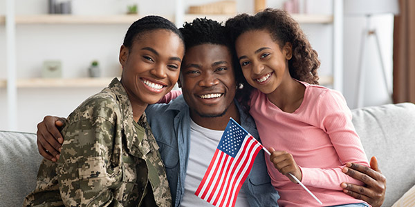 A Black woman in a military uniform sits smiling with a Black man and a little girl in their family home