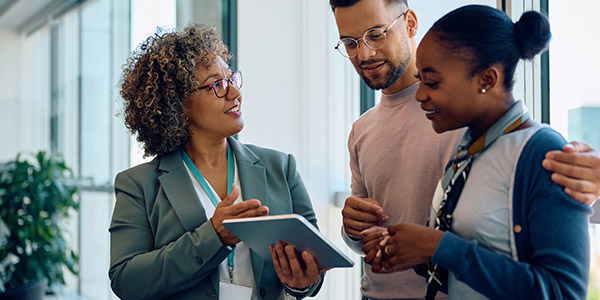 A Black woman dressed in a suite goes over something on a tablet with a smiling Black couple inside of a home
