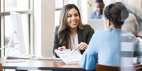 A woman dressed in a suit hands another woman a clipboard with paperwork on it