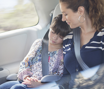 Mati in hči se vozita na zadnji klopi v avtomobilu. Hčerka spi z glavo na mamini rami, saj obe varuje varnostni pas.
