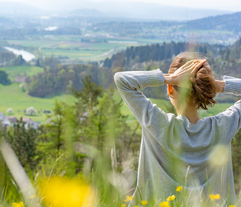 Mlada ženska sedi na trati, obkrožena s travniškimi cvetlicami, in uživa v spomladanskem soncu in lepem razgledu