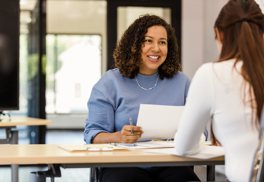 Two women sit across from each other at a table in an office. One woman is smiling and holding papers, while the other has her back to the camera. They appear to be having a professional conversation or interview.