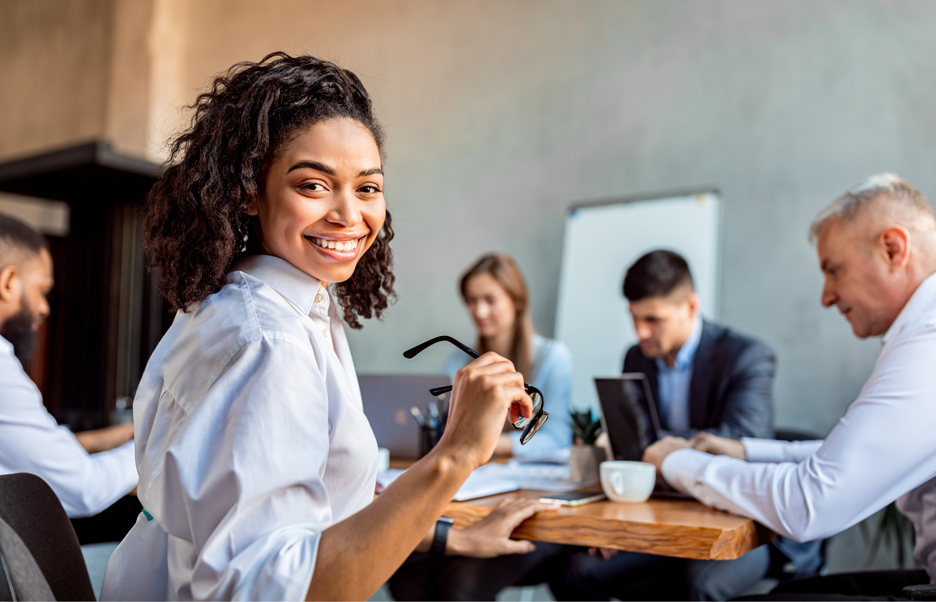 Smiling businesswoman holding glasses and looking at the camera during a team meeting in a modern office.
