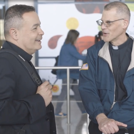 Two priests laugh outside of the airport, sporting Warriors to Lourdes apparel.