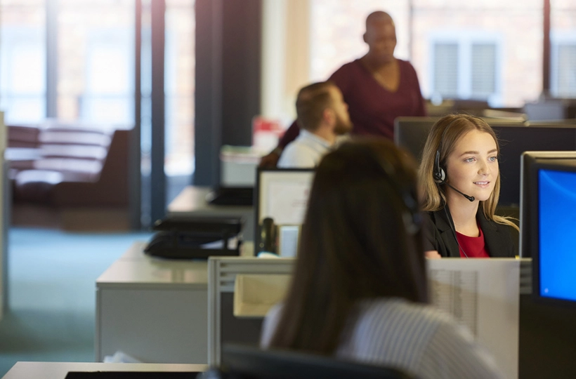 Photo of female Contact centre support worker with headset on at her desk talking to someone. Office environment