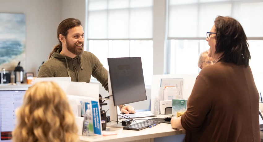 Two people interacting at an office reception desk, with a computer monitor, paperwork, and office supplies visible on the counter. 