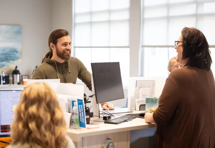 Two people interacting at an office reception desk, with a computer monitor, paperwork, and office supplies visible on the counter. 
