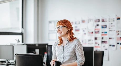 Young professional woman smiling holding coffee mug leaning against office desk 