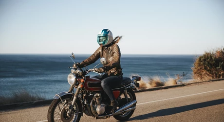 Female riding vintage motorcycle on lakeside paved road. A large body of water is in the background.  