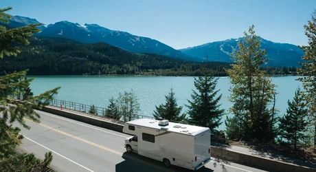 White RV traveling along a paved road beside a turquoise lake, with evergreen trees in the foreground and forested mountains in the distance under clear blue skies. 